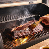 Whole brisket being basted with a thin dark sauce on a smoker grate with visible smoke