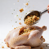 Atomic Chicken Chicken Stuffing 500g — spoonful of herbed stuffing mix frozen mid-drop into a raw chicken cavity