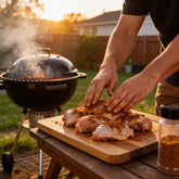 Atomic Chicken Jamaican Jerk Seasoning Rub 220g — man rubbing jerk seasoning onto raw chicken thighs beside the grill at sunset