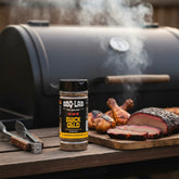 BBQ Lab Black Gold coffee & peanut butter BBQ rub — jar beside an offset smoker with smoked brisket and chicken