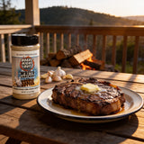 Boars Night Out Double Garlic Butter shaker beside a seared steak and roasted garlic on a porch table