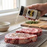 Chuds BBQ SPG 235g salt, pepper and garlic seasoning — a cook seasoning raw ribeye steaks from the shaker on a wire rack