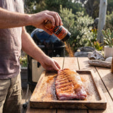 Coast n Smoke The Rip rub — pitmaster sprinkling the rub onto raw pork ribs at a backyard smoker
