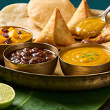 Goan Cuisine Chutney Trio bundle — spoonful of mango lime chutney lifted over a brass tray of three chutneys and samosas