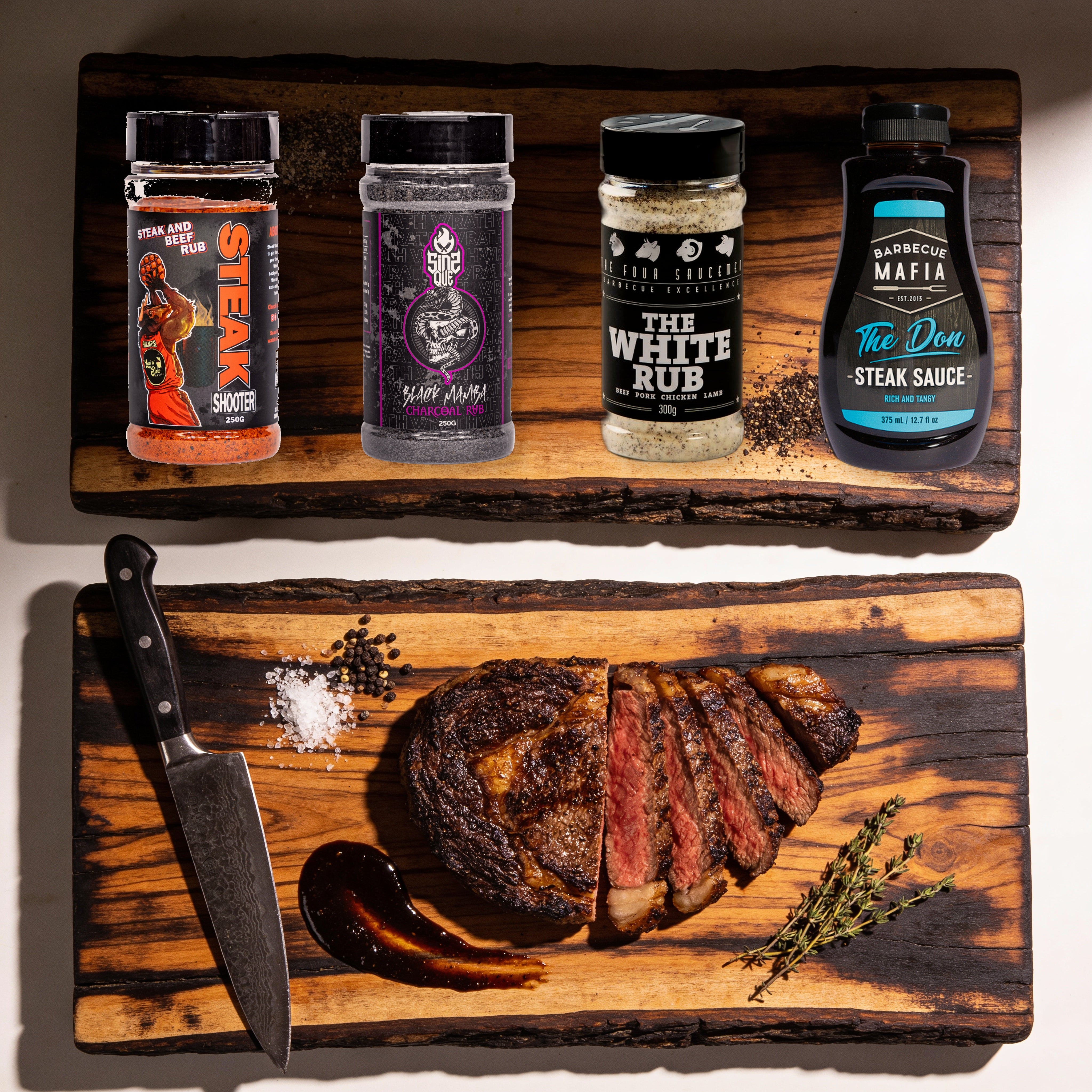 The Rub Hub High Steaks box - Steak Shooter, Black Mamba, The White Rub and The Don Steak Sauce on a timber board with a sliced scotch fillet