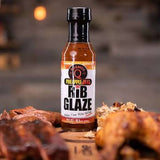 Kosmos Q Pineapple Heat Rib Glaze 439g glaze — bottle beside glazed pork ribs and barbecue chicken on a wooden board