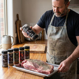 Middle Kingdom BBQ seasoning bundle 5 pack — home cook shaking seasoning over a raw beef brisket on a kitchen bench
