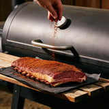 Seasoning a raw beef rib plate with Plowboys Bovine Bold Rub beside an offset smoker