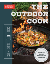 The Outdoor Cook by America's Test Kitchen paperback cookbook — front cover with paella over a fire pit