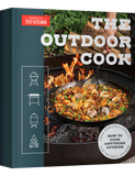 The Outdoor Cook by America's Test Kitchen paperback cookbook — front cover with paella over a fire pit