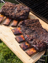 The Outdoor Cook by America's Test Kitchen paperback cookbook — smoked beef ribs sliced on a board