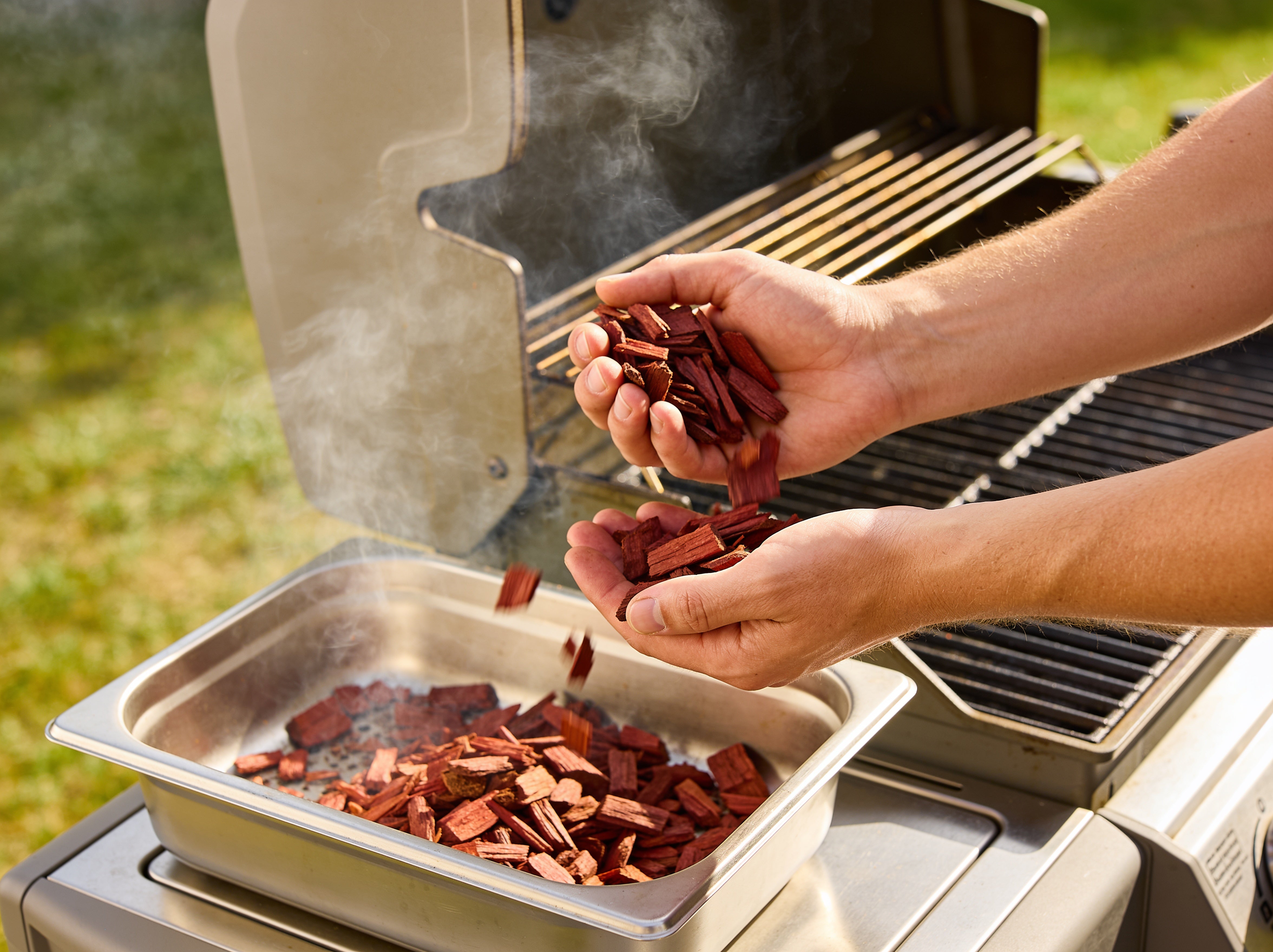 Western BBQ Products Cherry Smoking Wood Chips lifestyle photo — loading cherry wood chips into a smoker tray on the grill