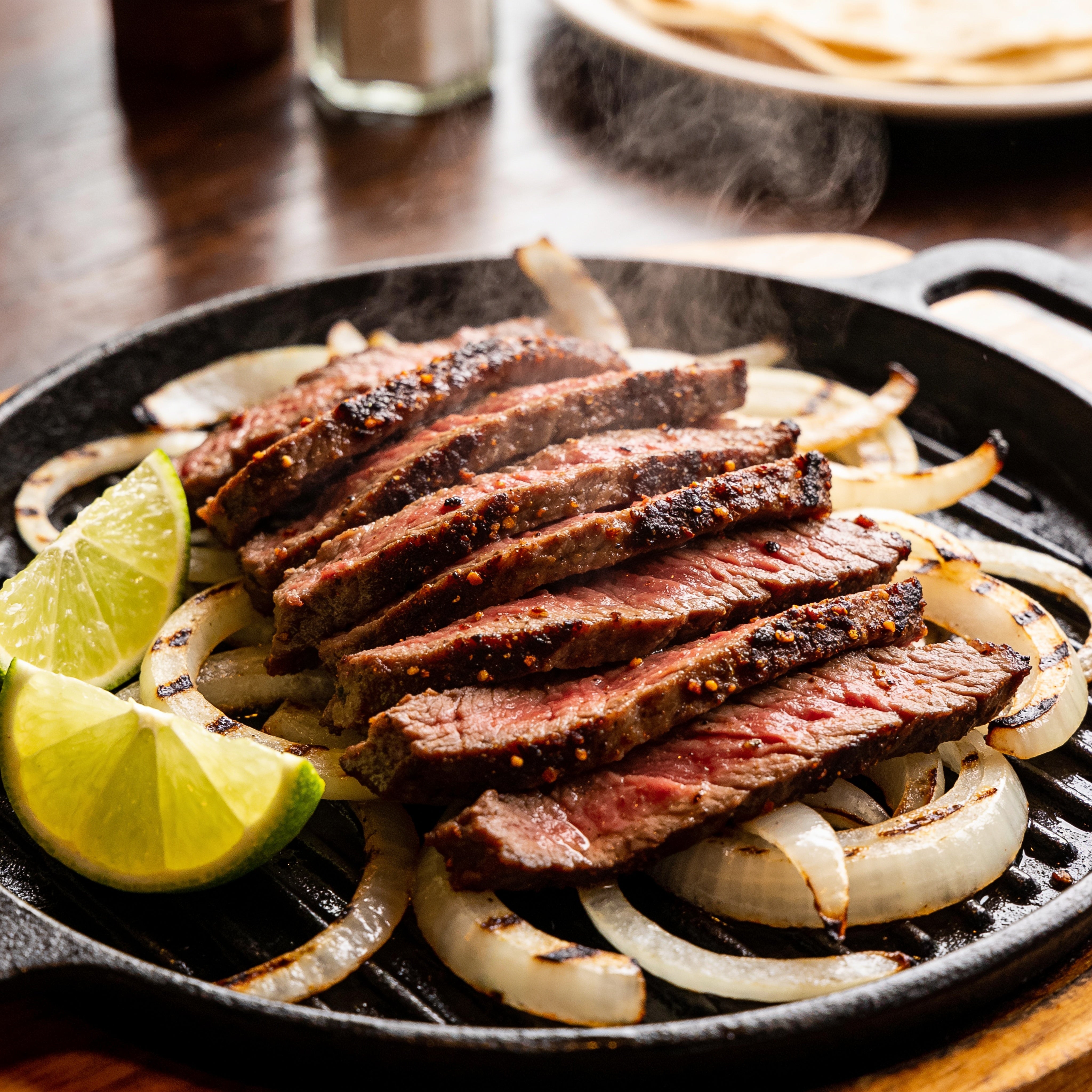 Sizzling skirt steak sliced for fajitas on a hot cast-iron platter with steam, lime wedges and charred onions