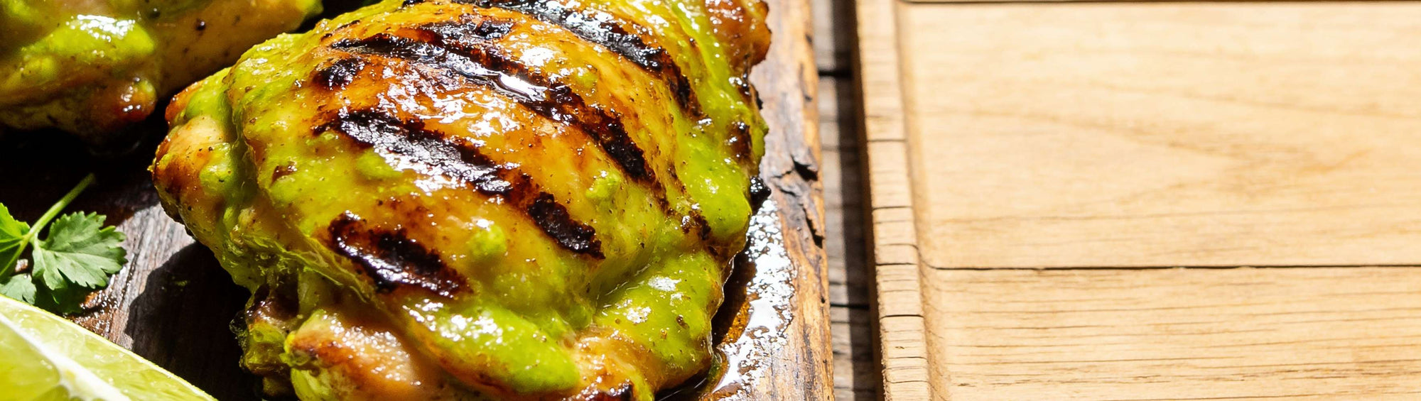 Verde-glazed grilled chicken thighs with lime wedges and cilantro on a dark rustic wood board