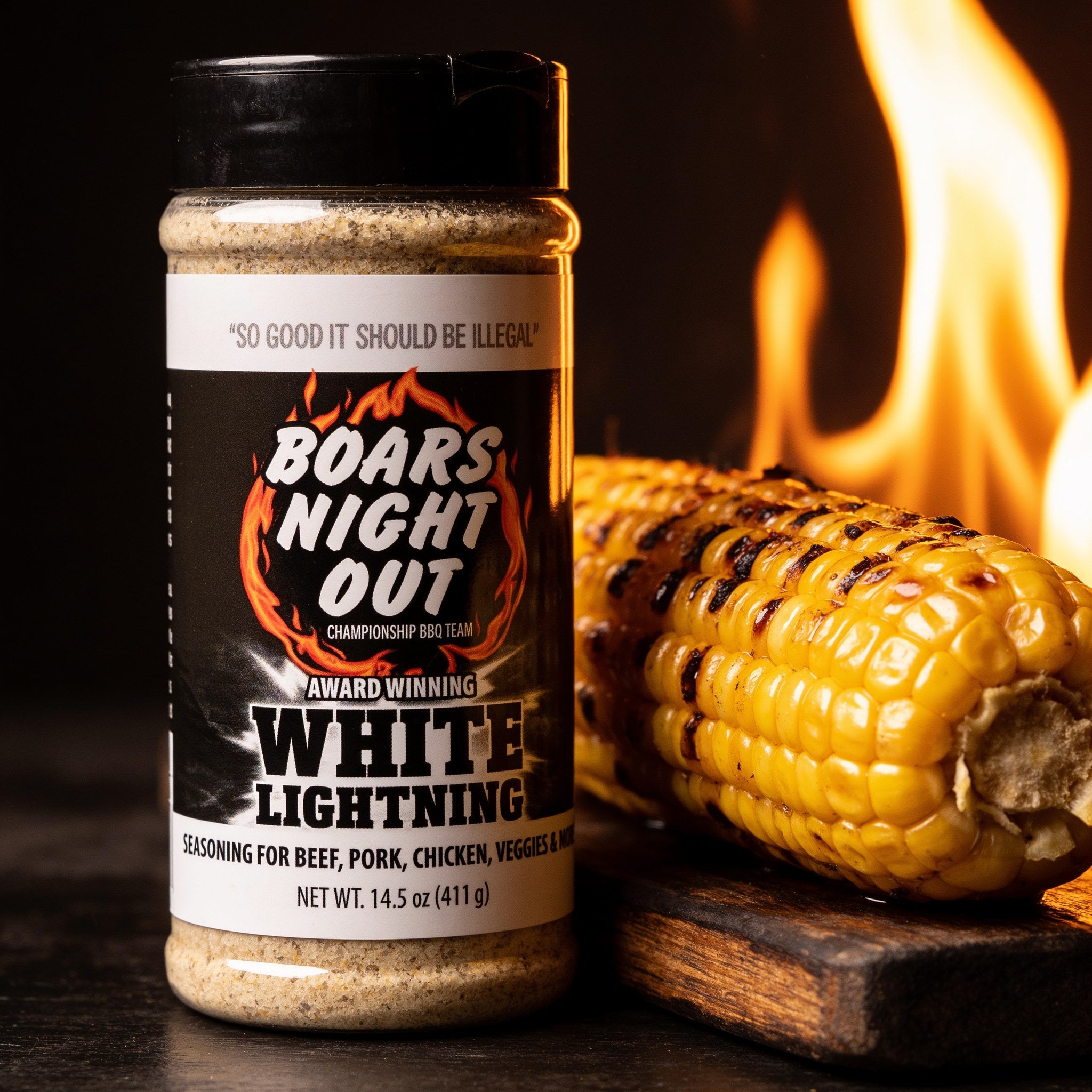 Close-up of charred grilled corn on the cob beside a Boars Night Out White Lightning shaker with fire glowing behind