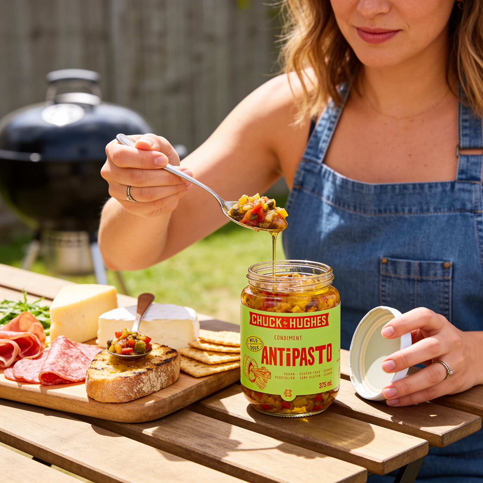 Chuck Hughes Mild Antipasto 375ml antipasto condiment — spooning antipasto from an open jar onto a backyard grazing board