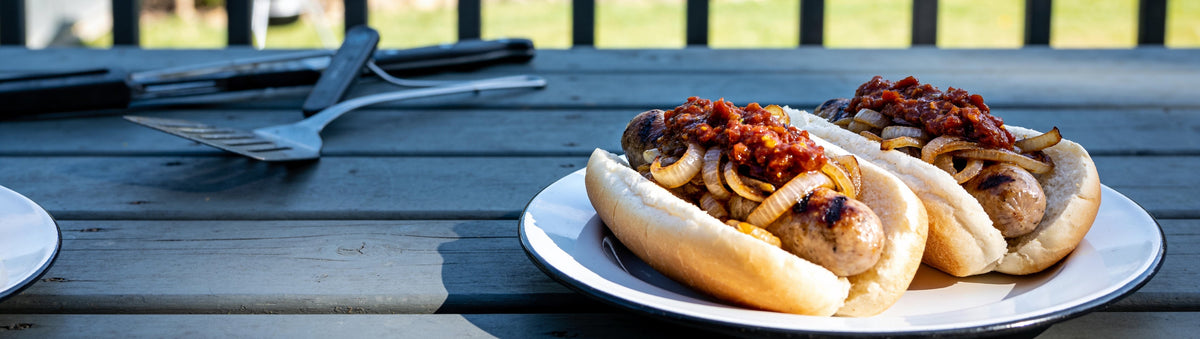 Grilled snags in rolls with onions and Cobra Chilli Hot Tamale Tomato Relish on a backyard deck