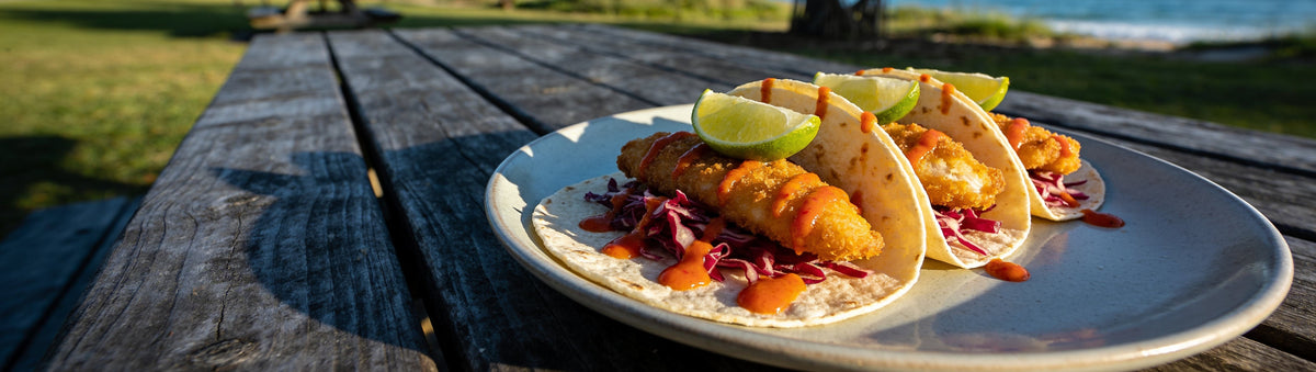 Crispy fish tacos drizzled with Cobra Chilli Summer Sizzle Chilli Sauce on a beachside picnic table