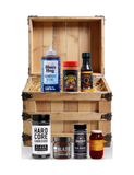 The Rub Hub The Quartermaster BBQ subscription box — example crate with Blues Hog Champions' Blend, Meat Church The Gospel, Kosmos Q, Hardcore Carnivore Black, BLEND Blazed honey, Big Bark and Proud Phoenix chilli crisp — contents change monthly, on transparent background