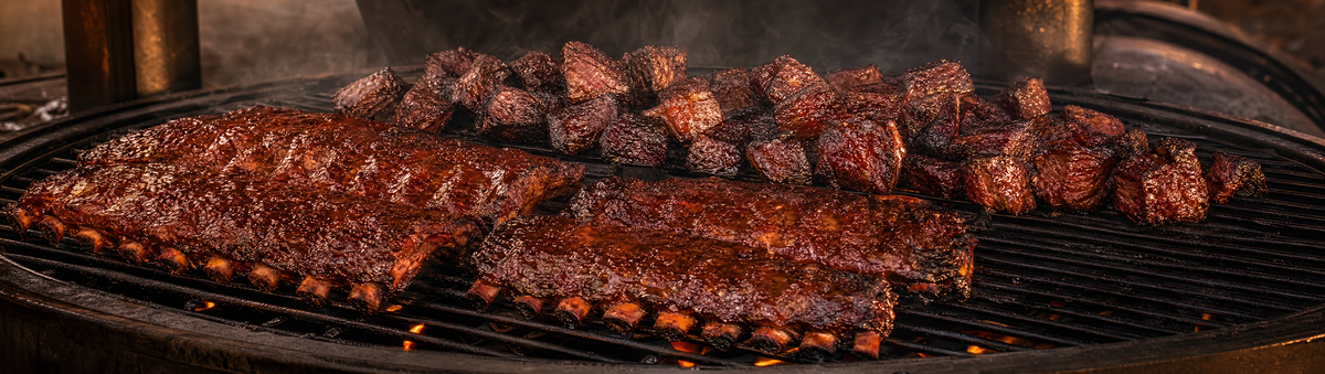 Glazed pork ribs and glossy burnt ends resting on a smoker grate at dusk