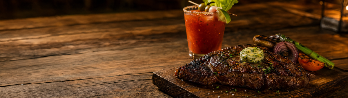 A marinated grilled steak resting on a wood board with a garnished cocktail nearby