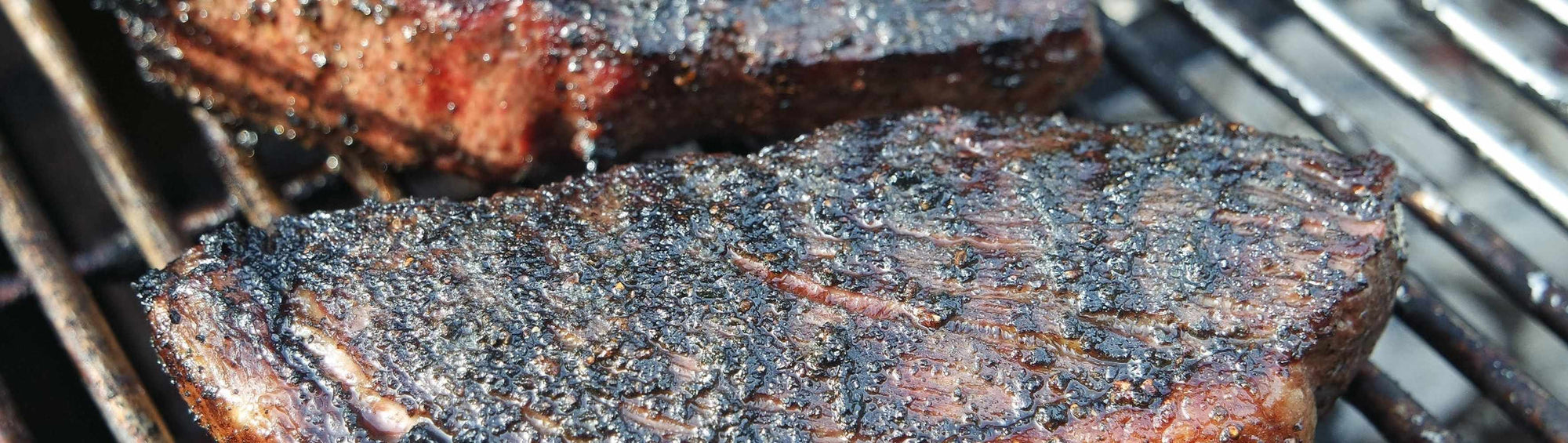 Hardcore Carnivore High Heat Gloves beside a seared beef cut with a dark peppery bark sizzling on a glowing charcoal grill