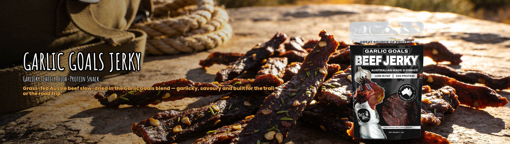 Low n Slow Basics Garlic Goals beef jerky banner with garlicky jerky strips on a rock outdoors