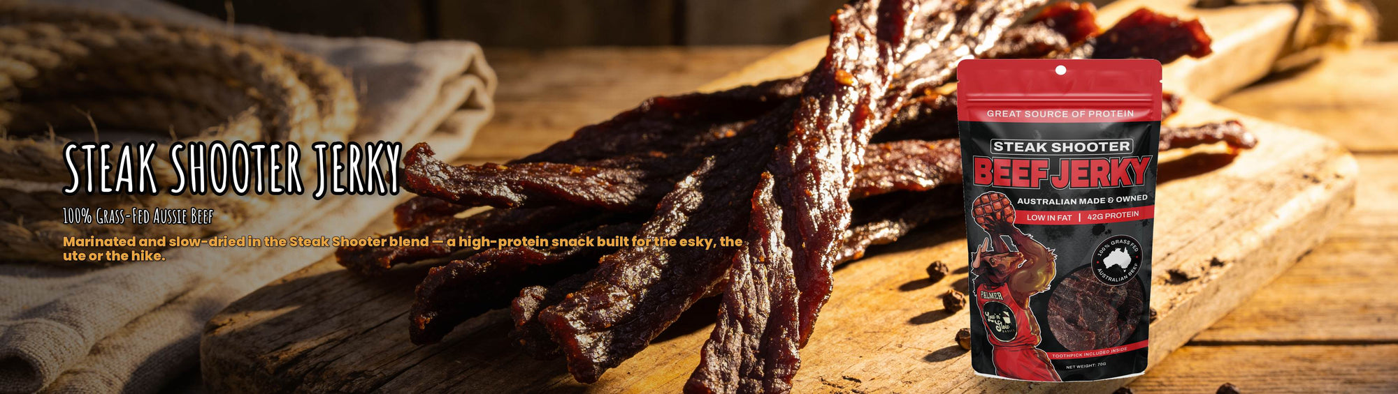 Low n Slow Basics Steak Shooter grass-fed beef jerky banner with dark glossy jerky strips on a rustic board