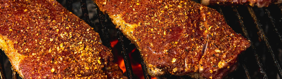 Hardcore Carnivore Steak Master Seasoning beside thick-cut steaks crusted in coarse seasoning searing over an open-flame grill