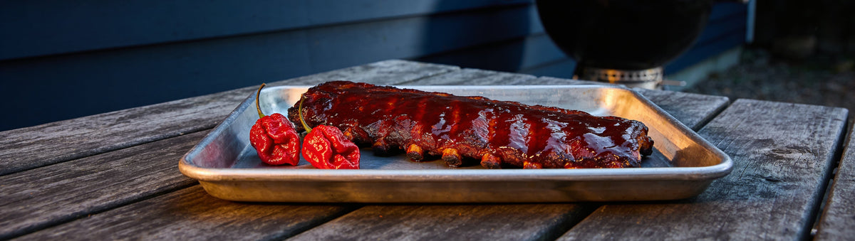 Glossy saucy pork ribs with two Carolina reaper chillies on a metal tray – Taipan BBQ Carolina BBQ Sauce