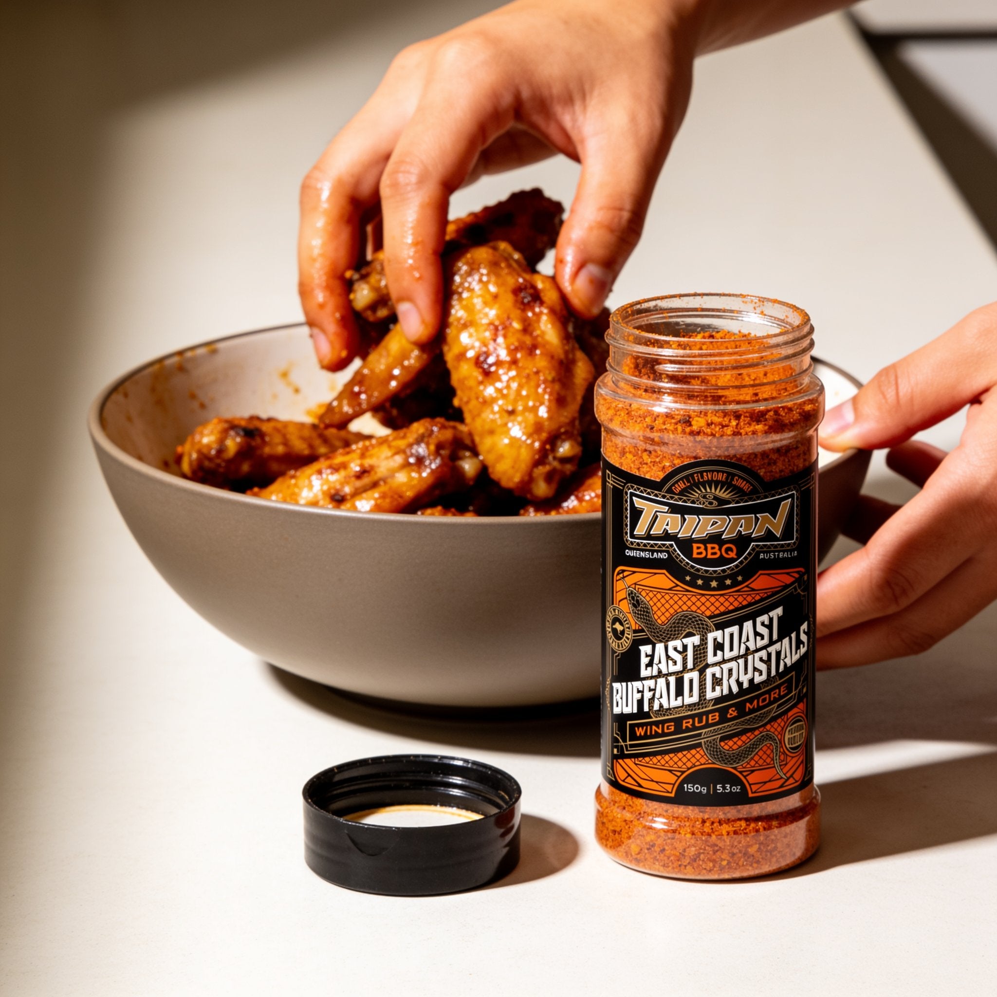 TAIPAN BBQ East Coast Buffalo Crystals buffalo wing seasoning 150g — use shot, hands tossing cooked wings with the seasoning in a bowl