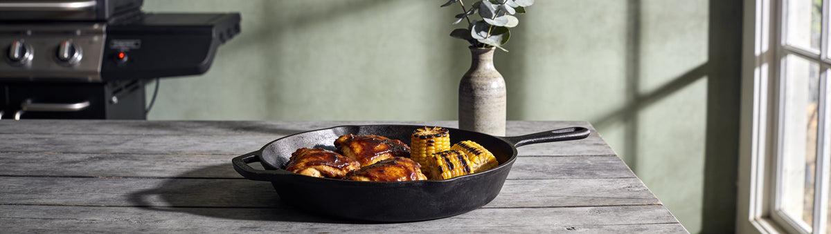 Sticky glazed chicken thighs and charred corn in cast iron on a grey timber table – Taipan BBQ Smoky Glaze