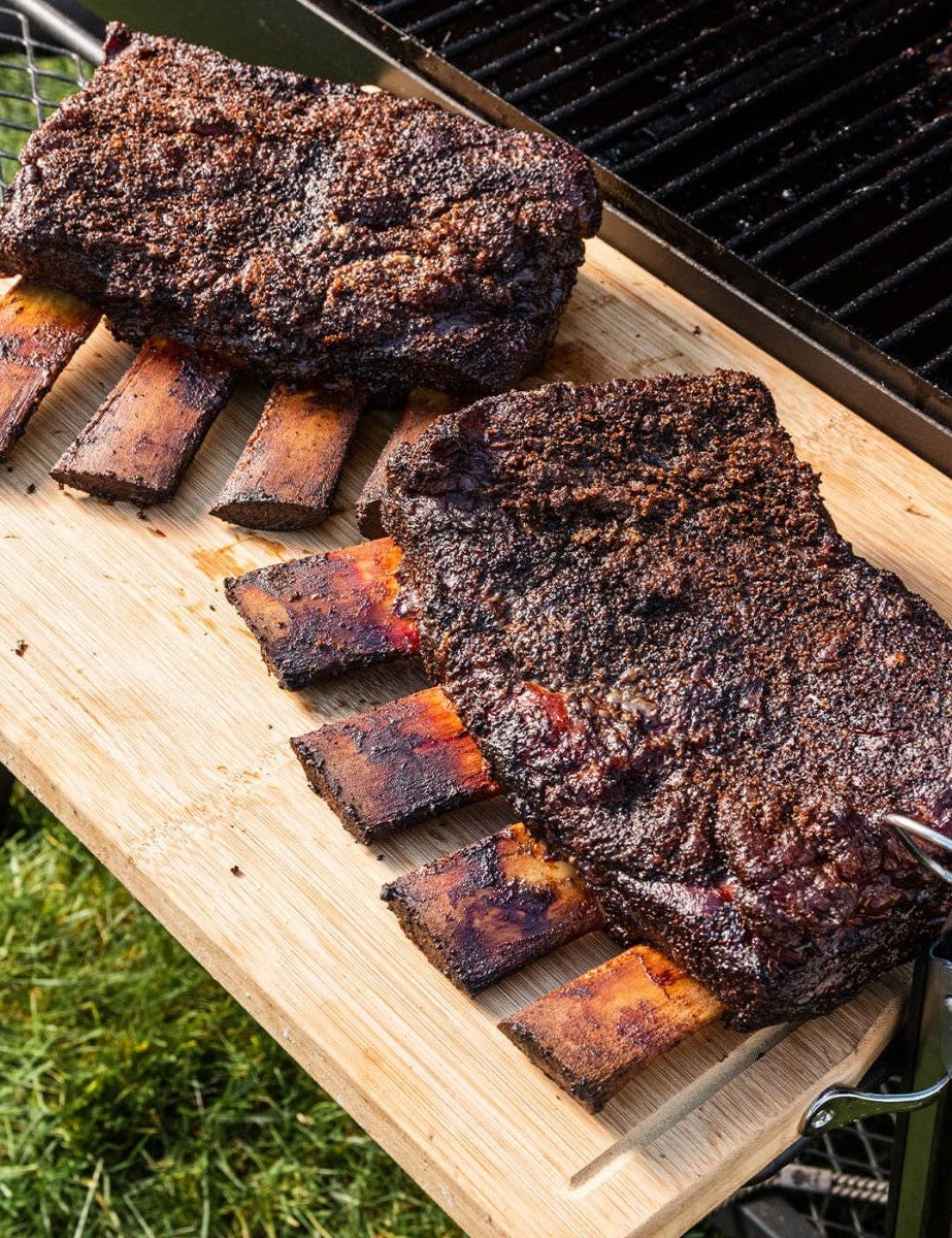 The Outdoor Cook by America's Test Kitchen paperback cookbook — smoked beef ribs sliced on a board